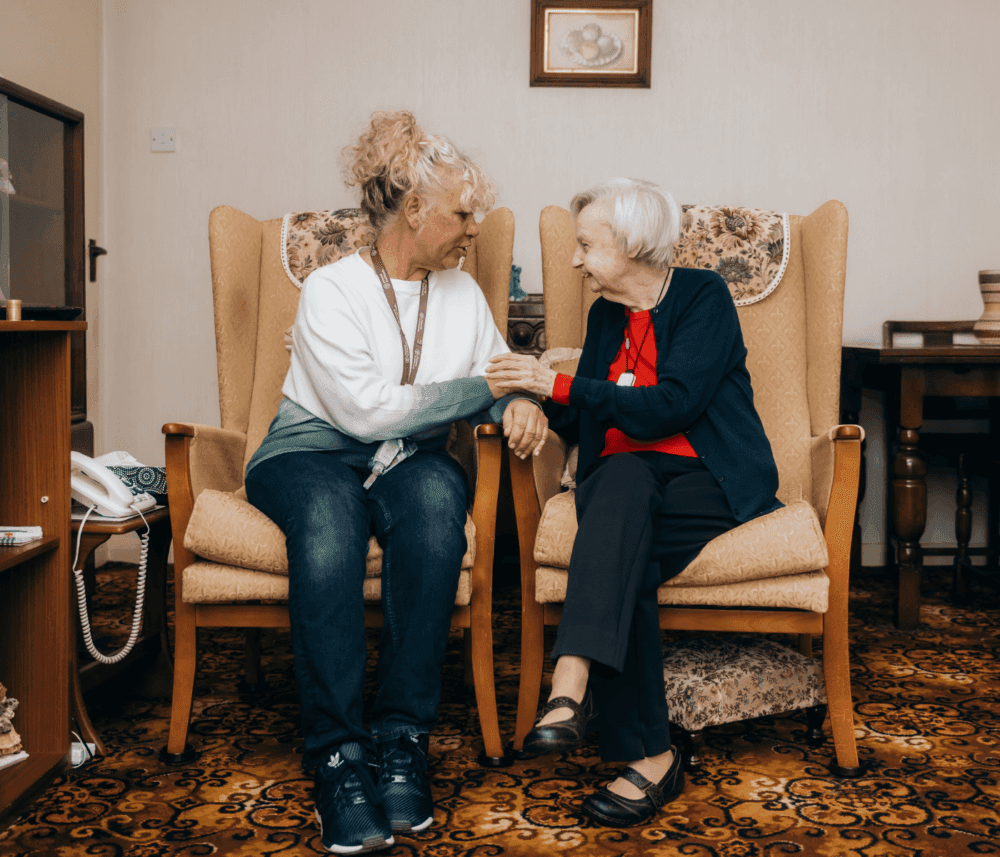 An elderly woman and a caregiver sit in armchairs, smiling and holding hands in a cozy living room. - Home Instead