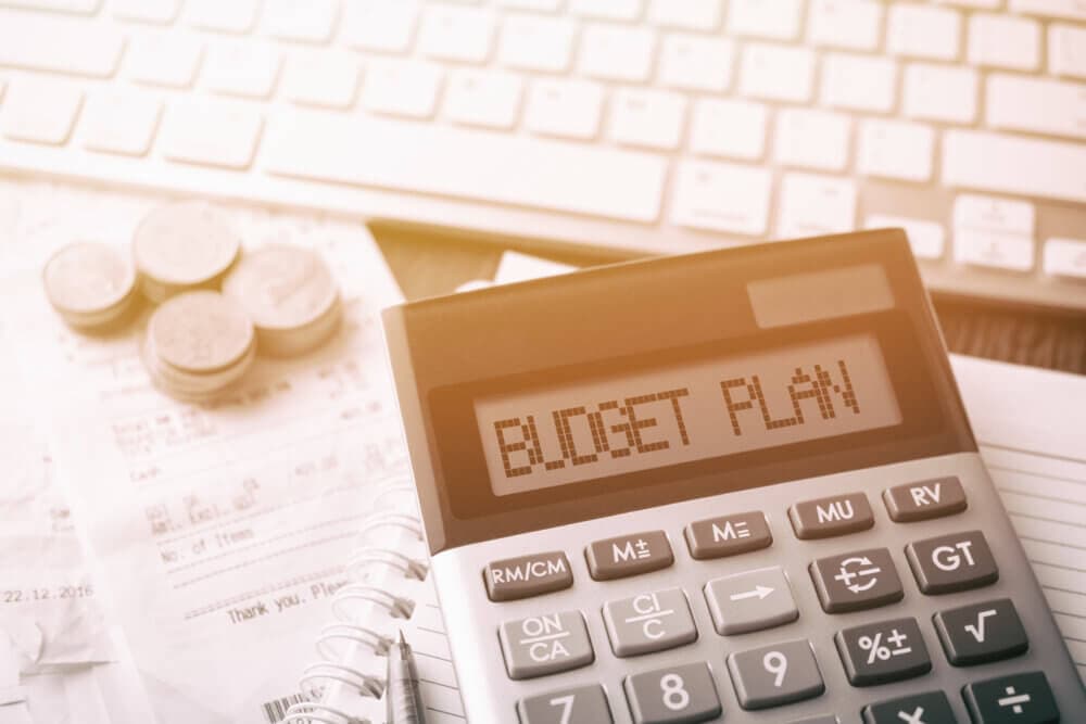 A calculator displaying "BUDGET PLAN" on its screen, with coins, receipts, and a keyboard in the background. - Home Instead
