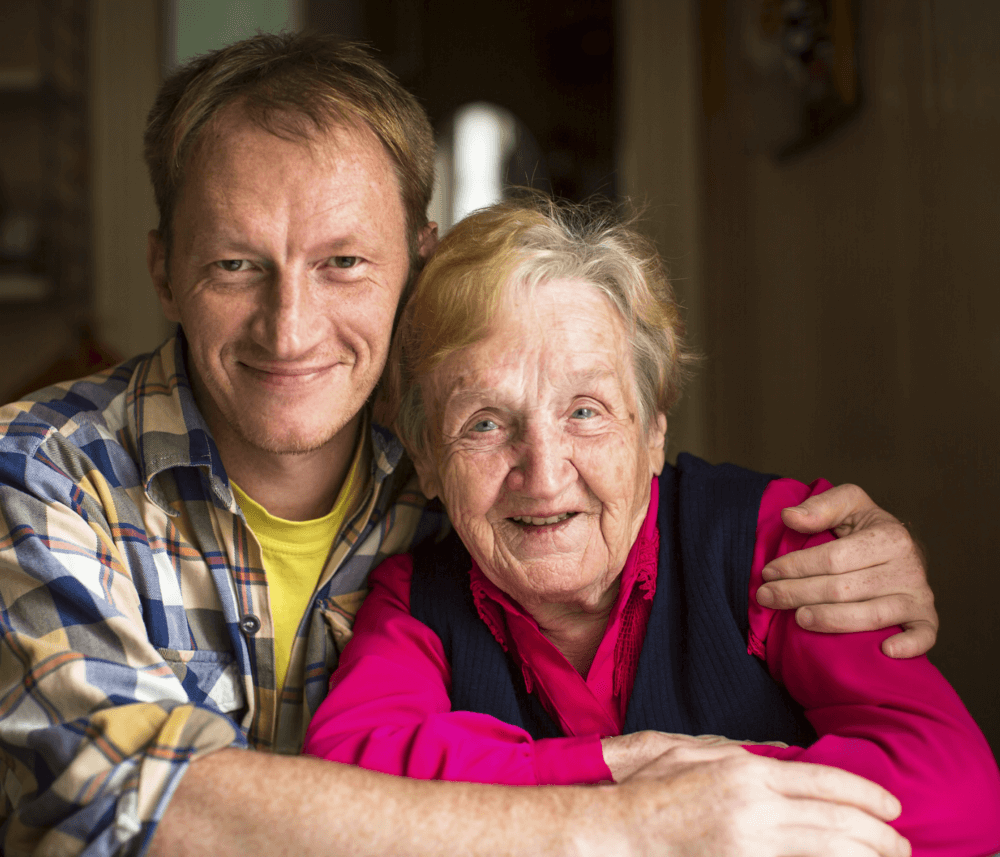 A smiling man with his arm around an elderly woman, both sitting closely and looking at the camera. - Home Instead