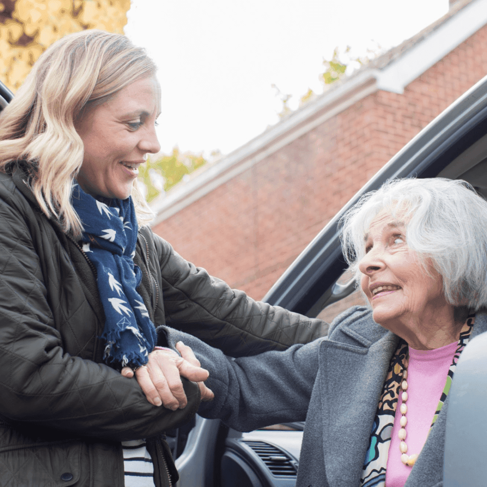 A younger woman helps an older woman out of a car while holding her hand, both smiling at each other warmly. - Home Instead