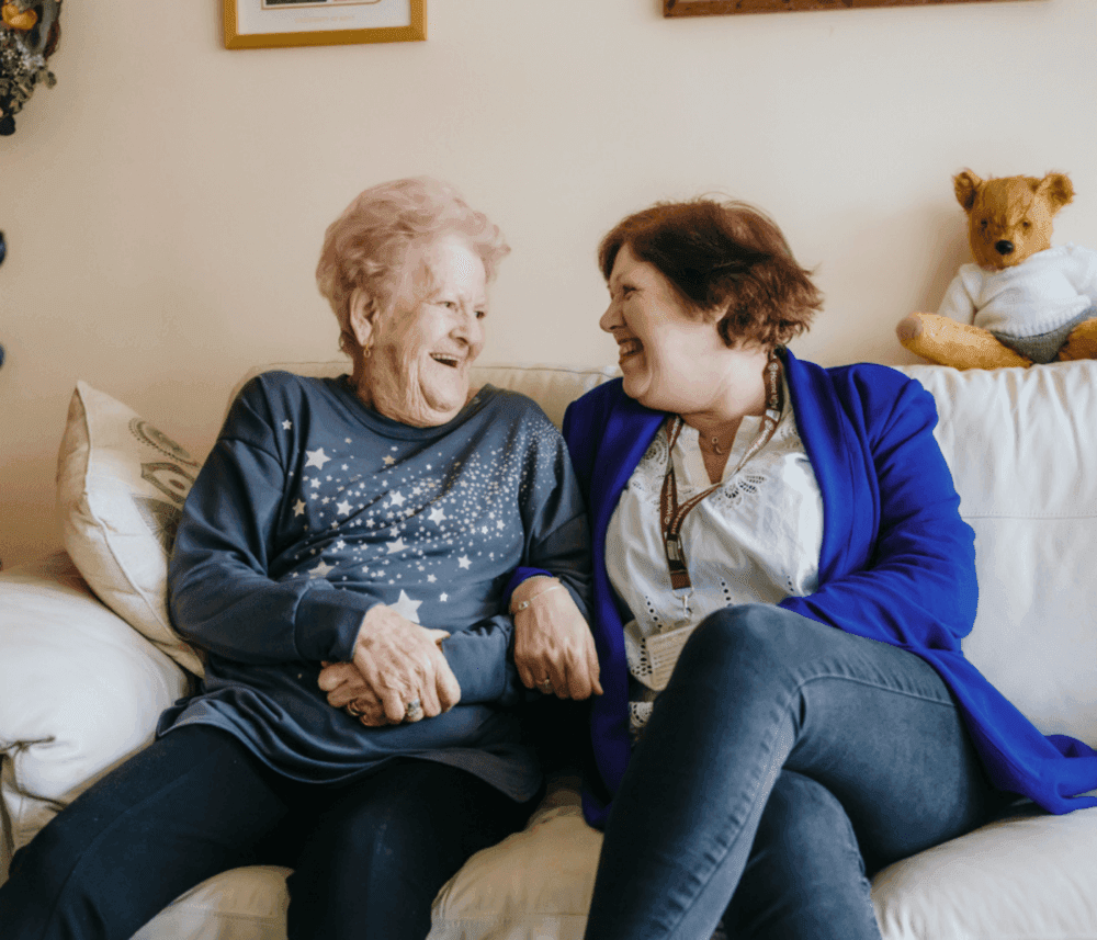 Two women, one elderly and one middle-aged, sit on a couch, laughing and holding hands in a warmly decorated room. - Home Instead