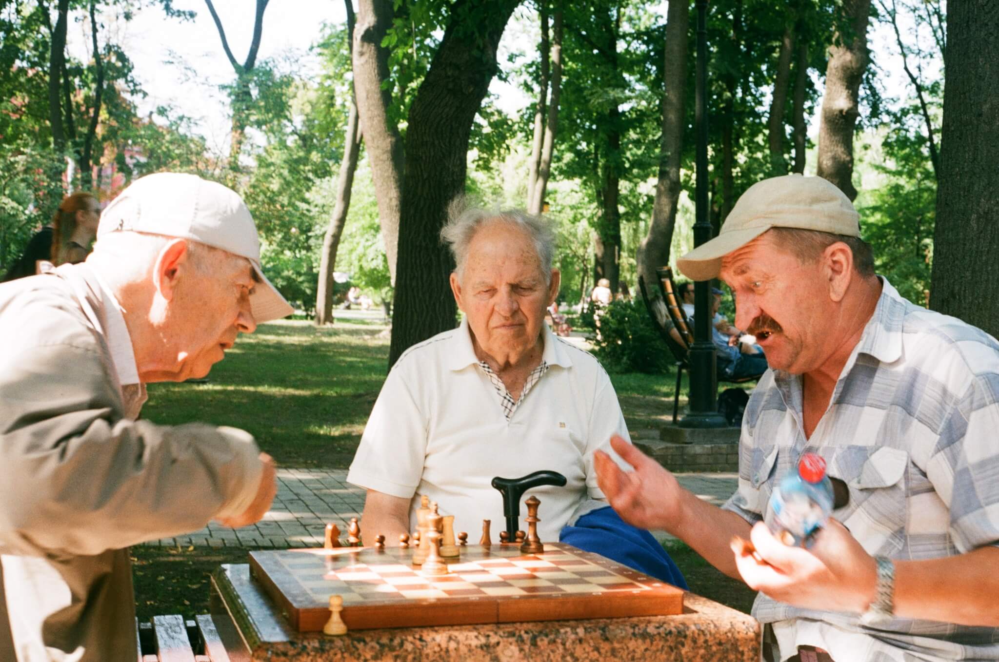 Three elderly men playing chess at a park, with trees and people in the background. - Home Instead