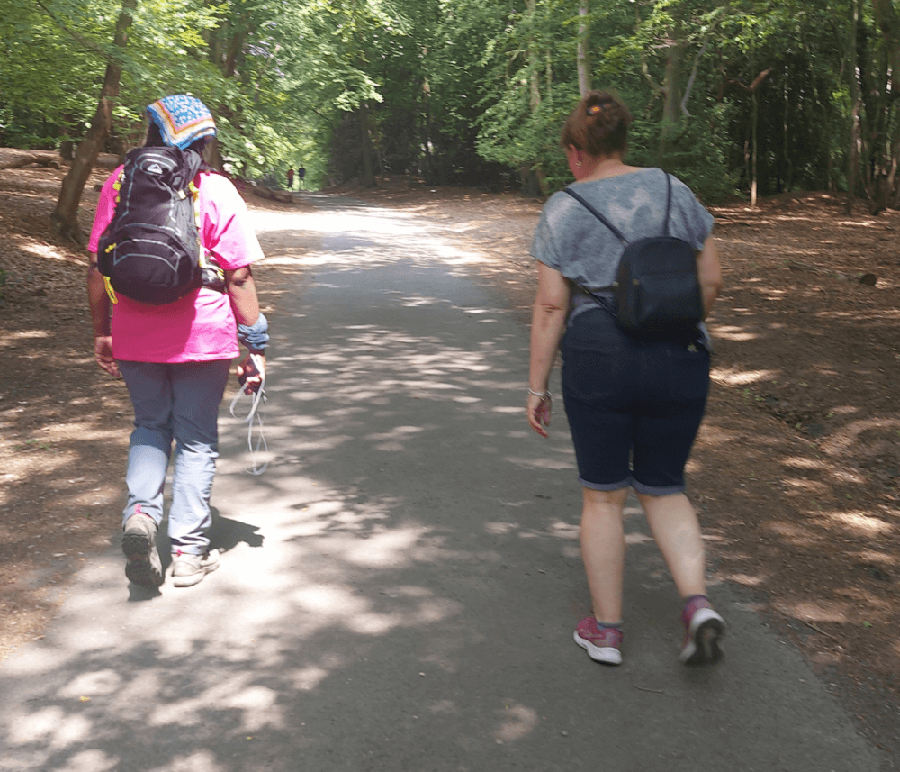 Two people walking on a forest path, wearing casual clothes and backpacks, with trees lining the trail. - Home Instead