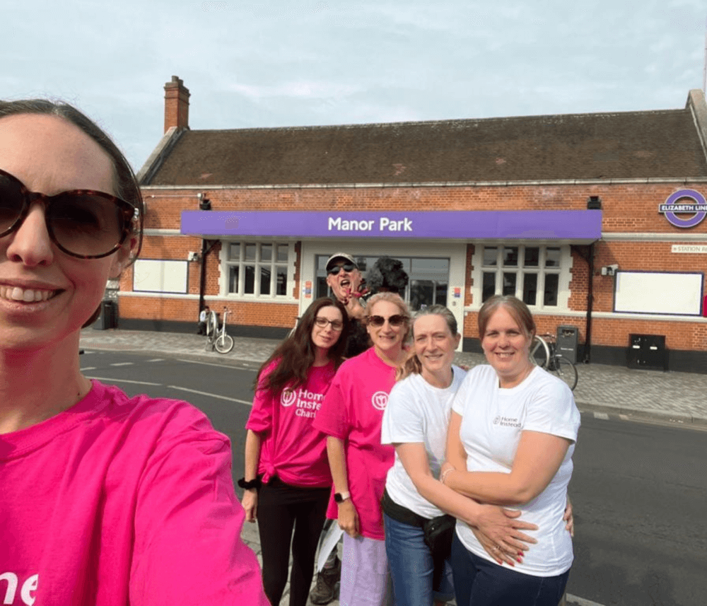 Group of people in pink and white shirts smiling outside Manor Park train station on a sunny day. - Home Instead