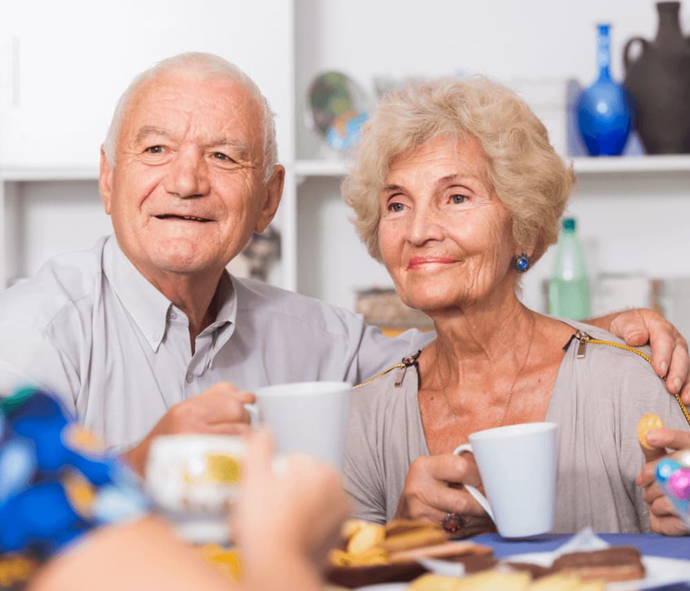 Elderly couple smiling and holding cups of tea at a table with snacks and other people visible in the foreground. - Home Instead