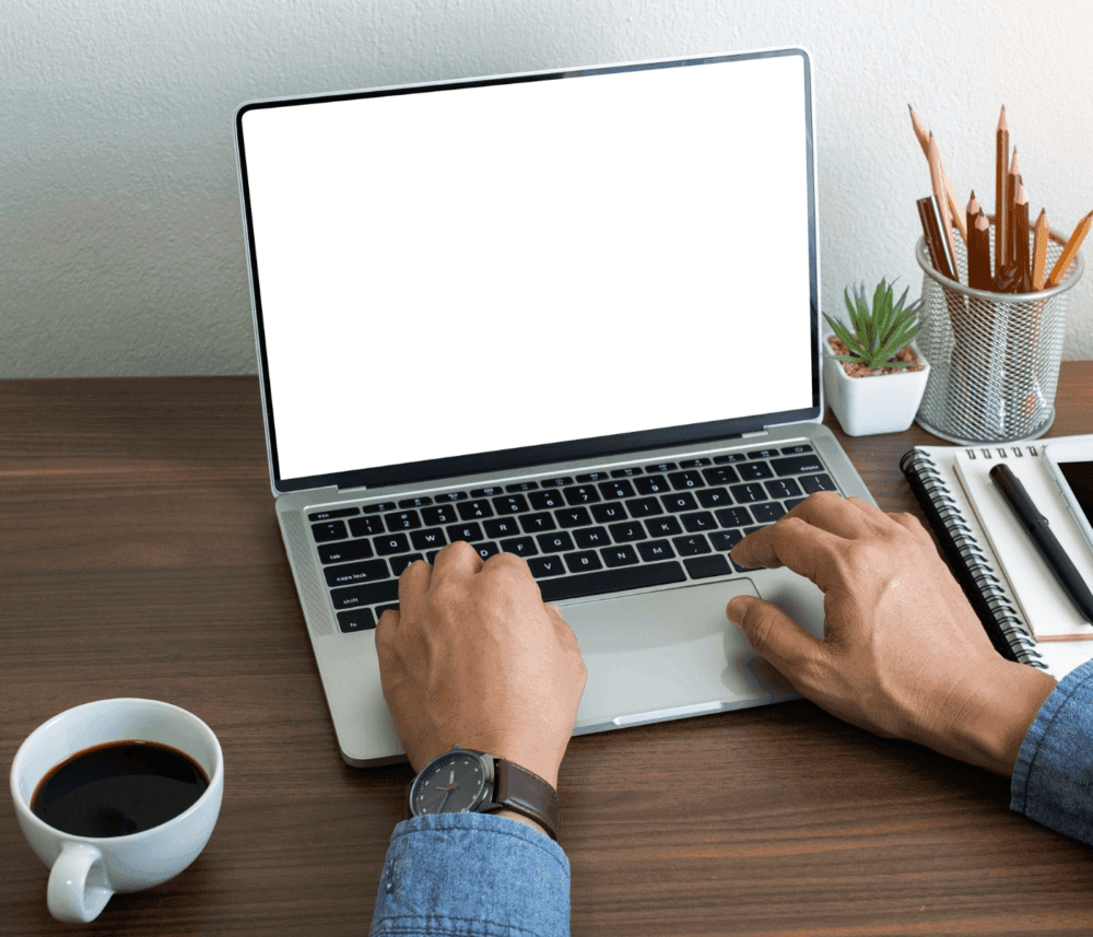 Person typing on a laptop with blank screen, a cup of coffee, notebook, and plant on a wooden desk. - Home Instead