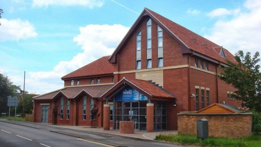 A modern red-brick building with steeply sloped roofs and large windows, featuring the signage "well" at the entrance. - Home Instead