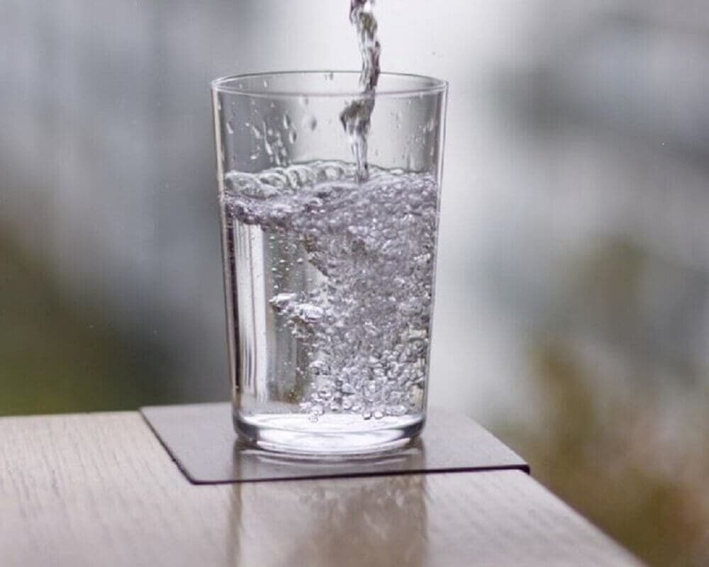 A clear glass being filled with water, placed on a coaster on a wooden surface, with a blurred background. - Home Instead