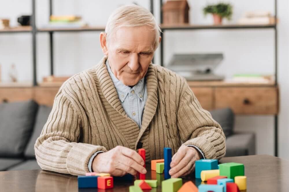 Elderly man focused on stacking colorful wooden blocks at a table, with shelves in the background. - Home Instead