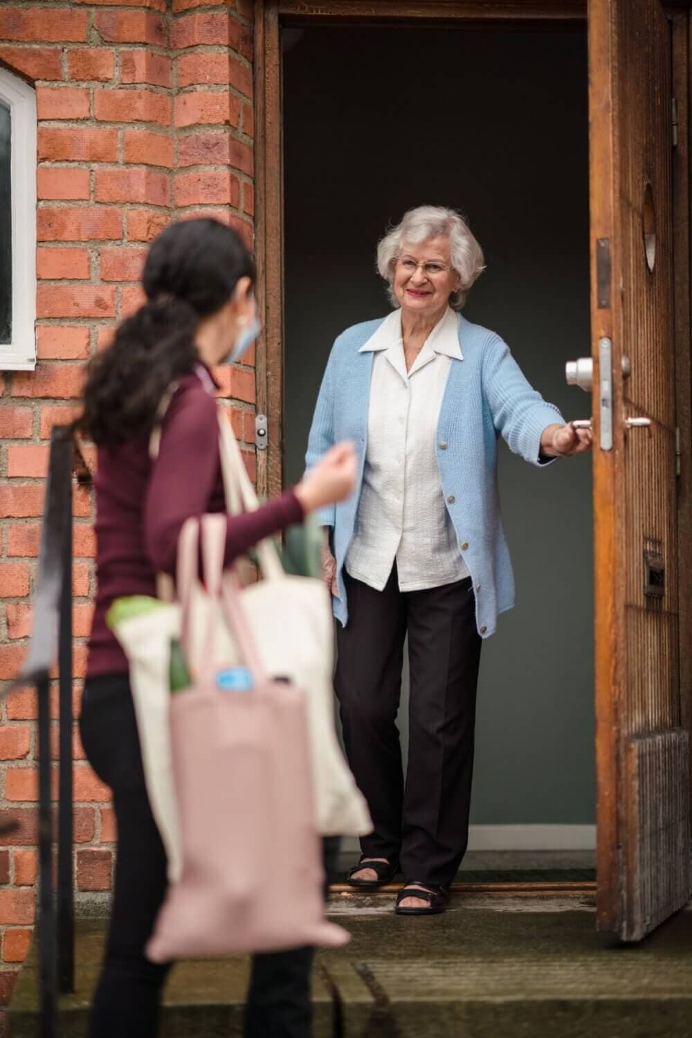 A senior woman standing at an open door greets a younger woman carrying tote bags on a doorstep. - Home Instead