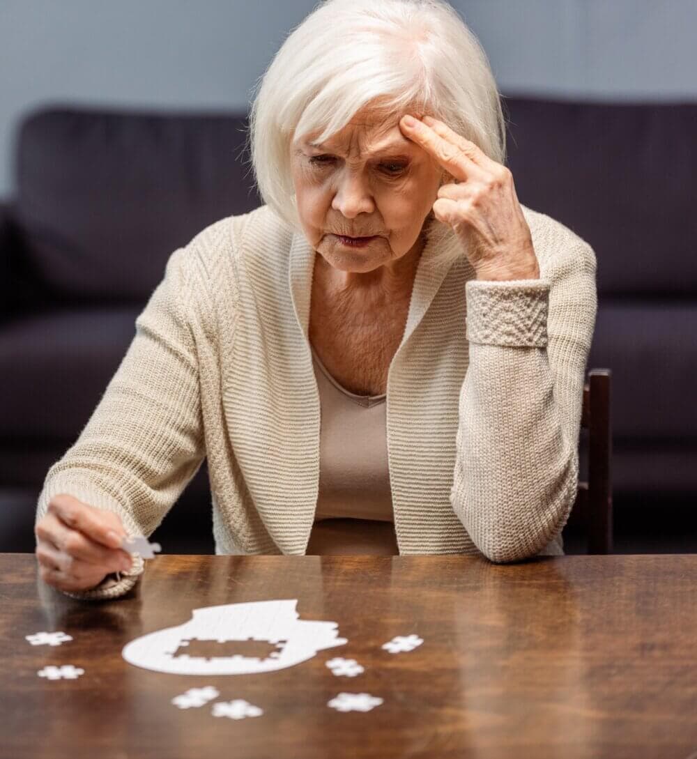 Elderly woman in a beige cardigan contemplating a jigsaw puzzle at a wooden table, hand to forehead in thought. - Home Instead