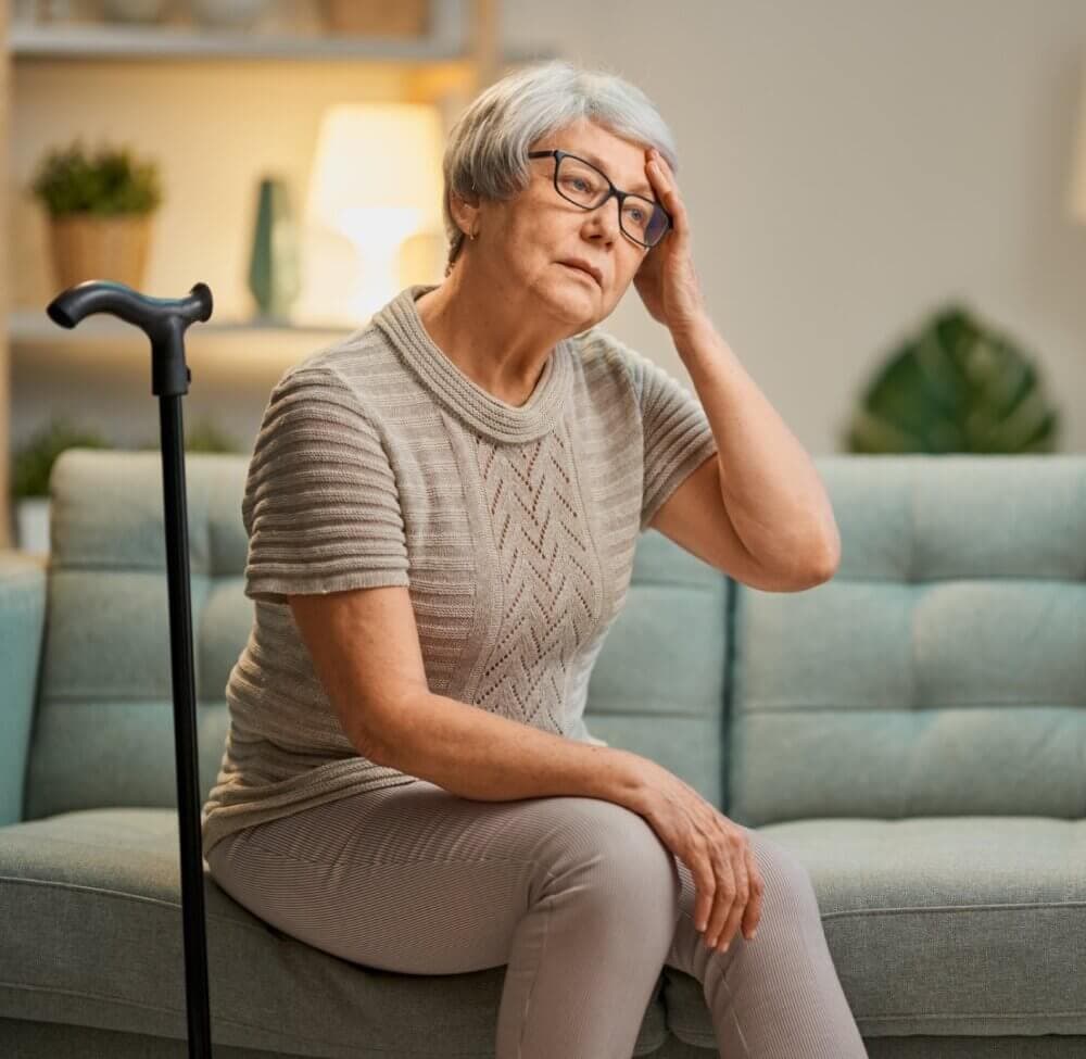 Elderly woman with glasses sits on a couch, leaning on her cane and resting her hand on her forehead, looking pensive. - Home Instead