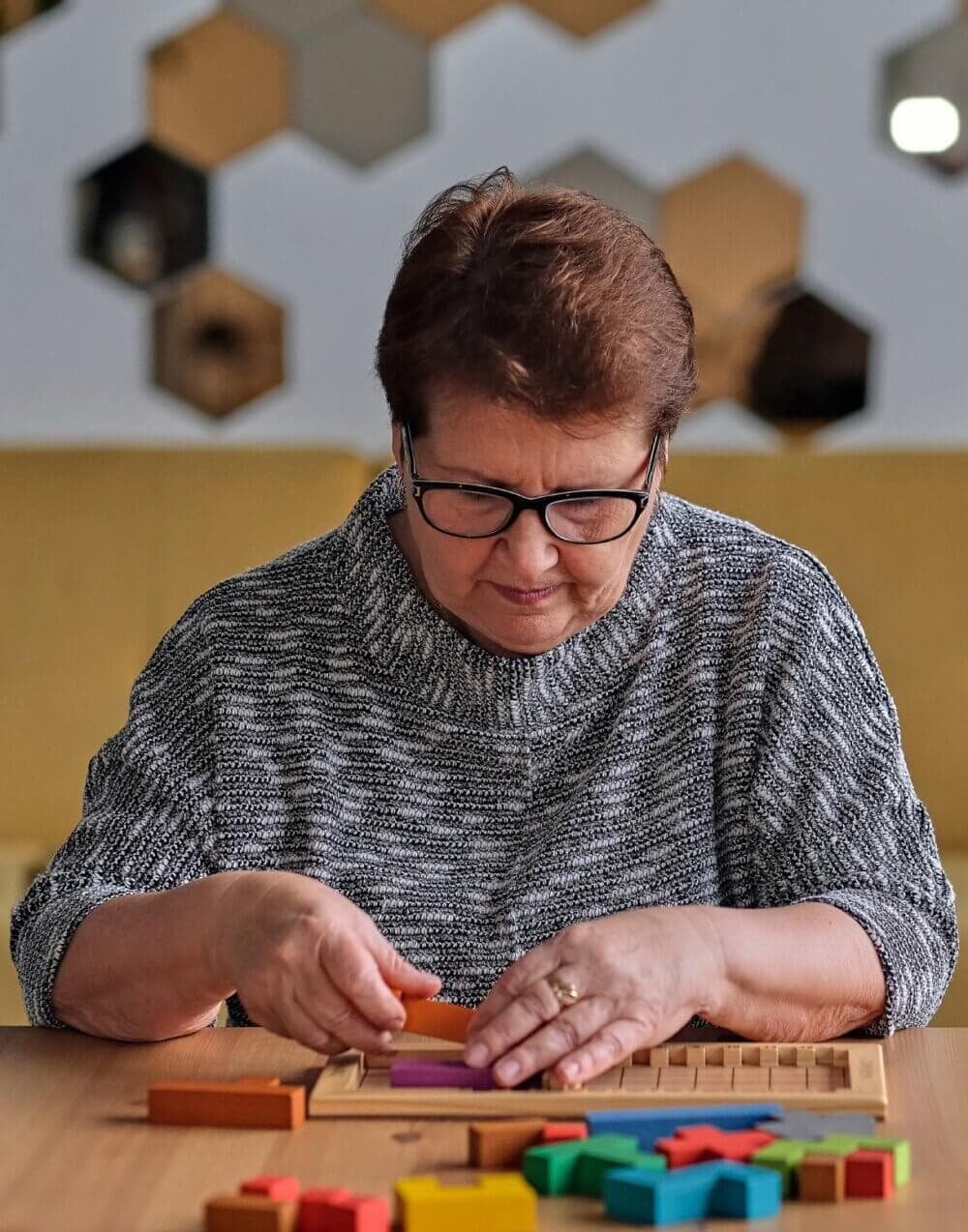 An elderly woman with glasses carefully arranges colorful wooden blocks on a table, focused on the task. - Home Instead