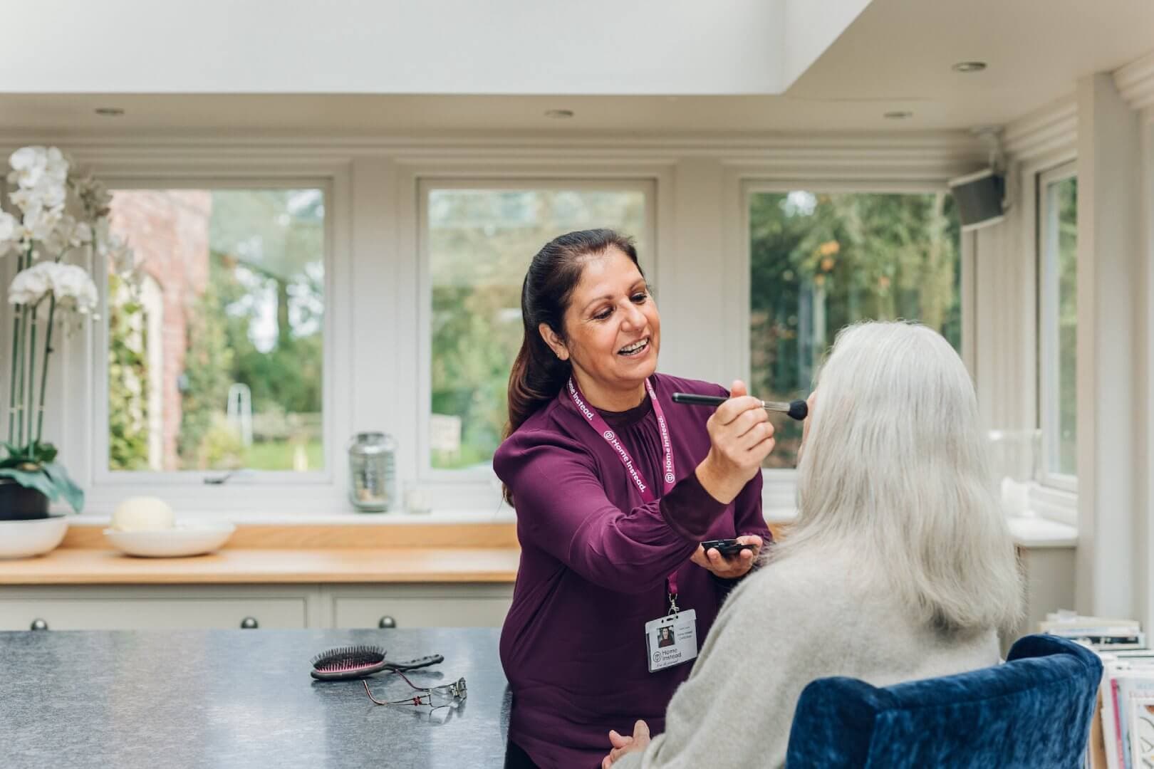 A woman wearing a name tag applies makeup to an older woman in a bright room with large windows. - Home Instead