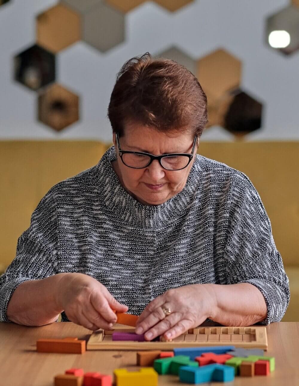 An elderly woman with glasses focuses on a wooden puzzle with colorful pieces at a table. - Home Instead