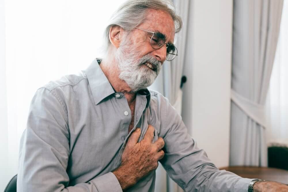 Elderly man with glasses clutching his chest in pain, sitting indoors by a window with curtains. - Home Instead
