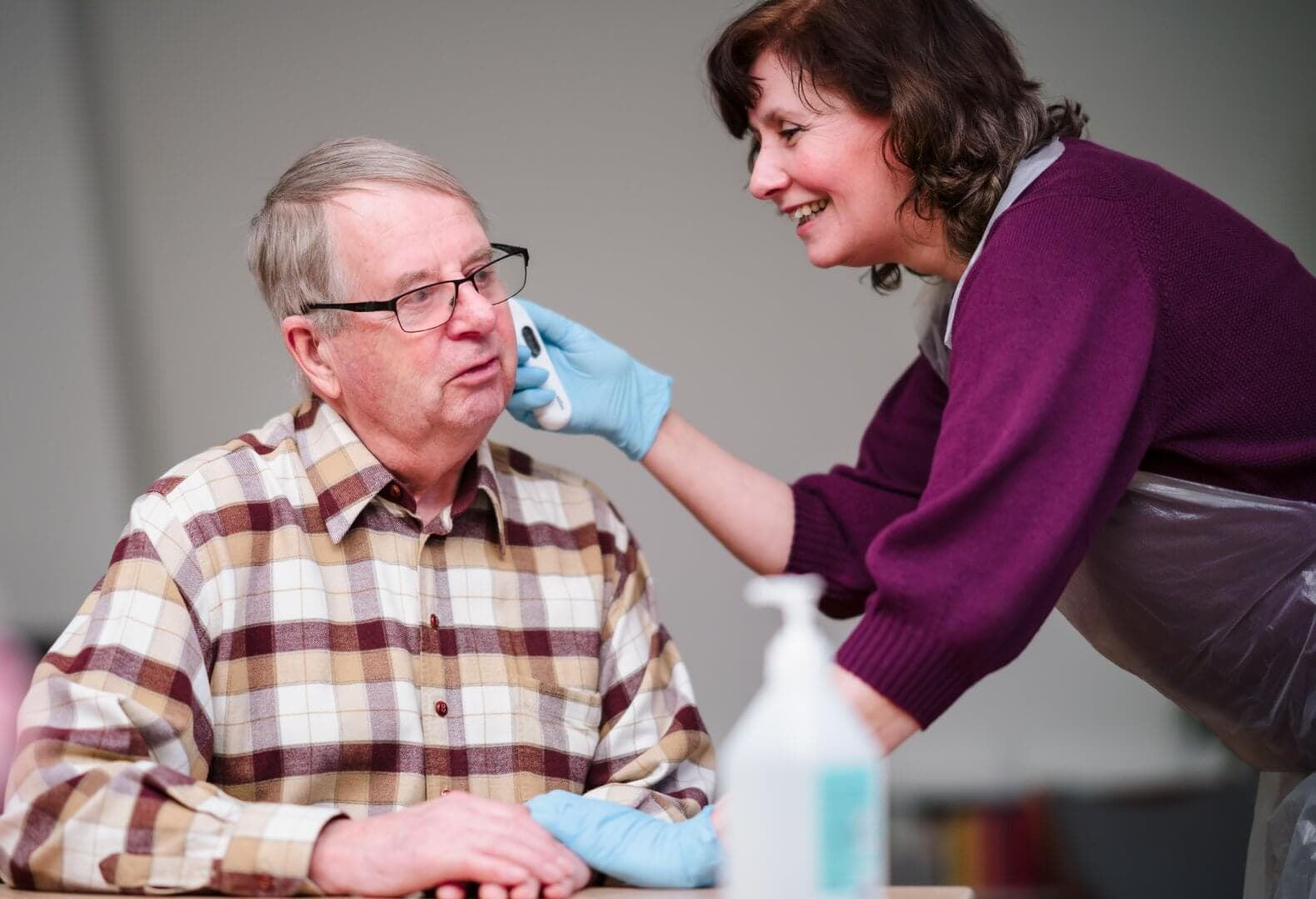 Woman in gloves and apron wipes elderly man's face as he sits at a table, both smiling and interacting warmly. - Home Instead