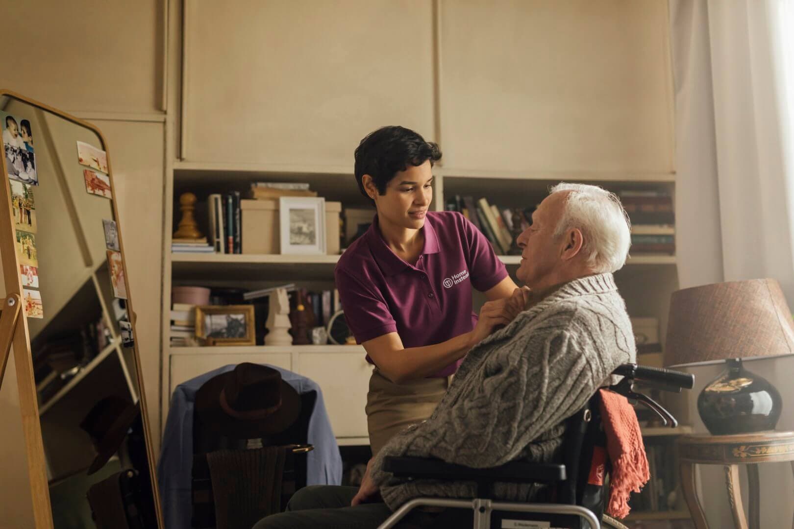 A caregiver in a purple shirt assists an elderly man in a wheelchair inside a cozy room with a bookshelf and mirror. - Home Instead
