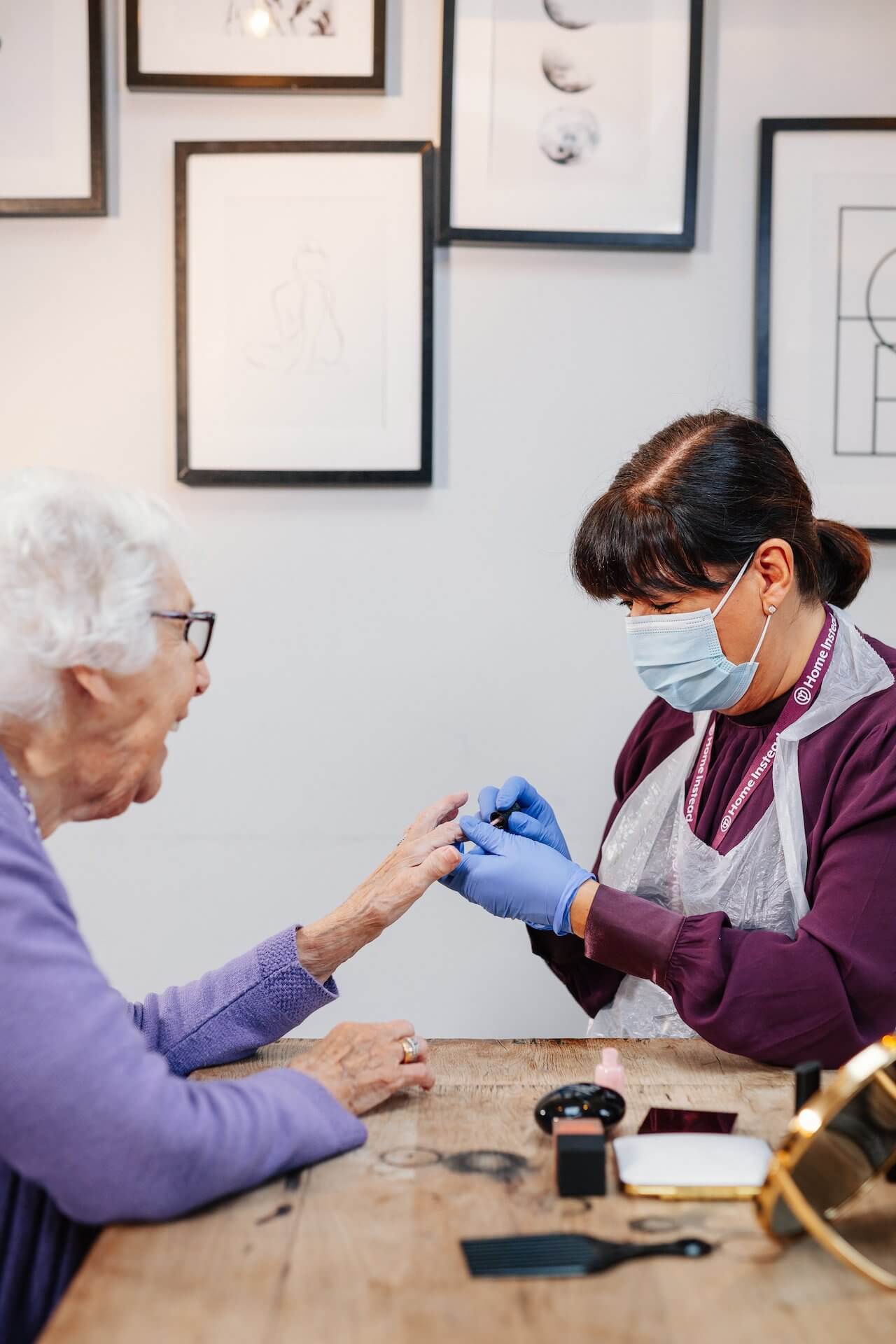 A nurse in PPE tends to an elderly woman's fingernails at a wooden table in a room with framed wall art. - Home Instead Southampton