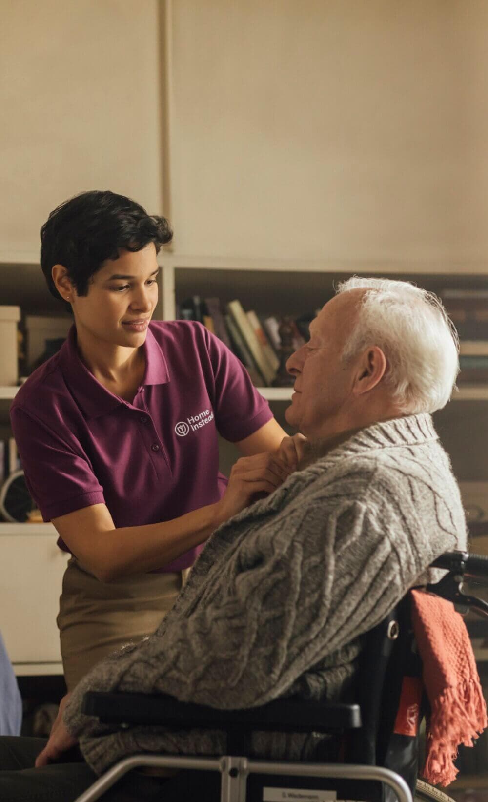 A caregiver in a purple shirt helps an elderly man in a wheelchair by adjusting his sweater. - Home Instead