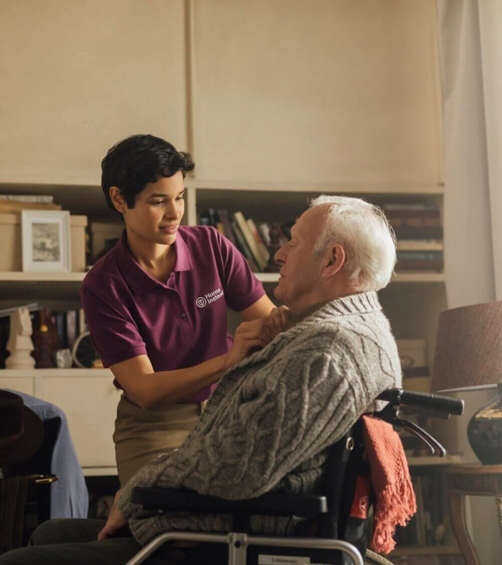 A caregiver in a purple shirt assists an elderly man in a wheelchair, adjusting his clothing in a cozy room. - Home Instead