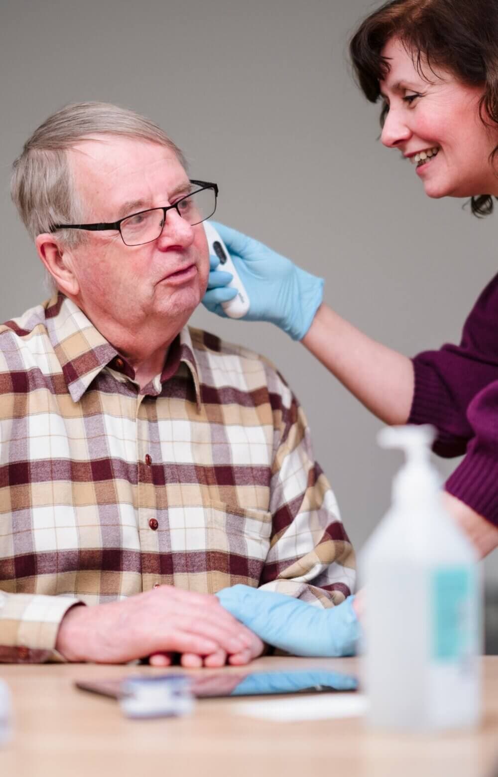 A healthcare worker in gloves wipes the face of an older man wearing glasses and a plaid shirt. - Home Instead