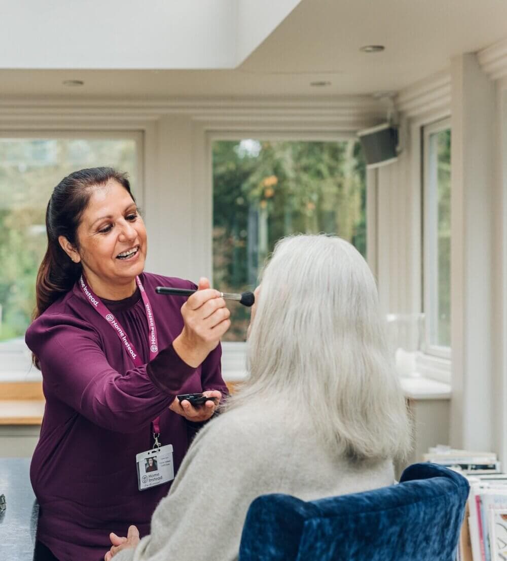 A woman with a name badge applies makeup to an elderly person with white hair, seated in a room with large windows. - Home Instead