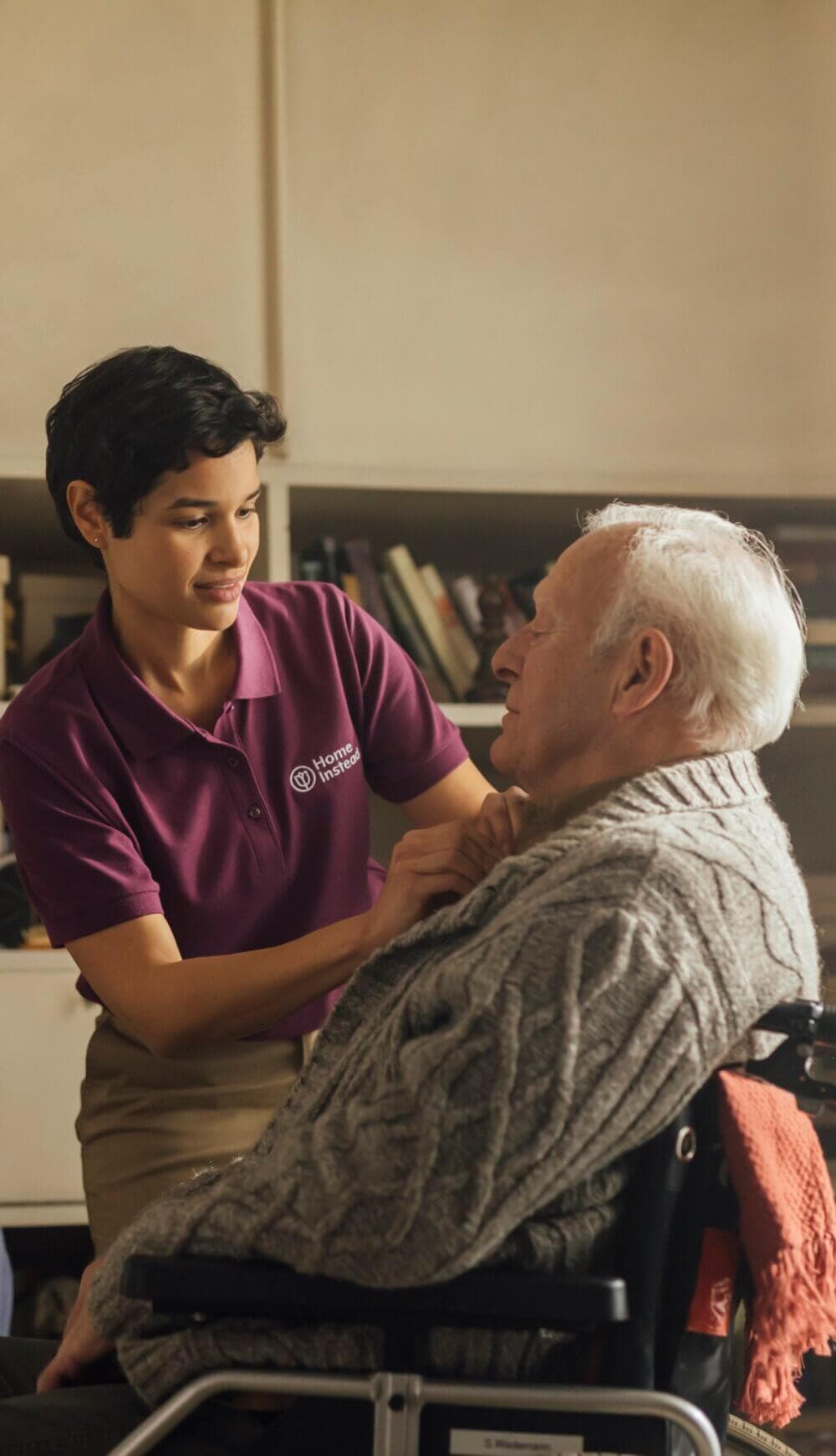 A caregiver helps an elderly man in a wheelchair with his sweater in a cozy, book-filled room. - Home Instead