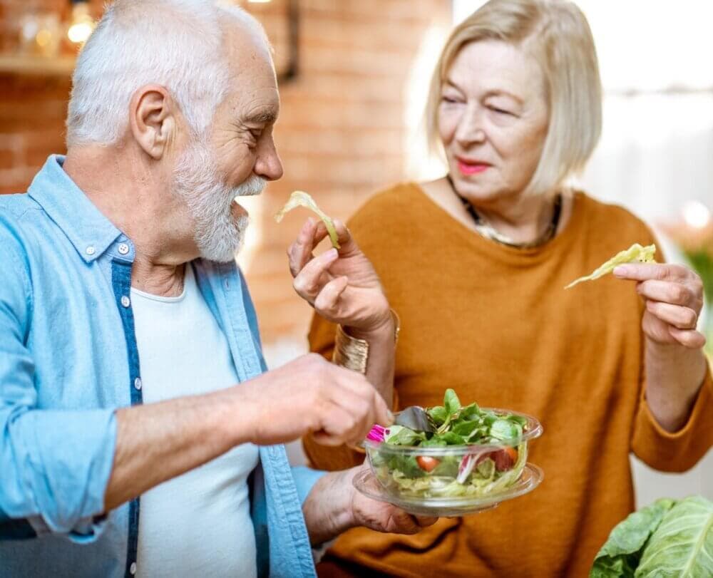 An older couple enjoys a salad together in a bright kitchen, smiling and holding pieces of lettuce. - Home Instead