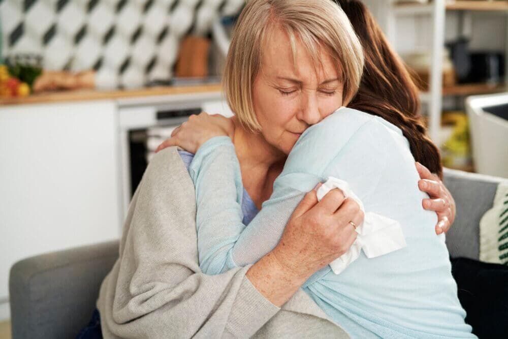 Elderly woman warmly hugging a younger woman in a kitchen, both showing emotional expressions. - Home Instead