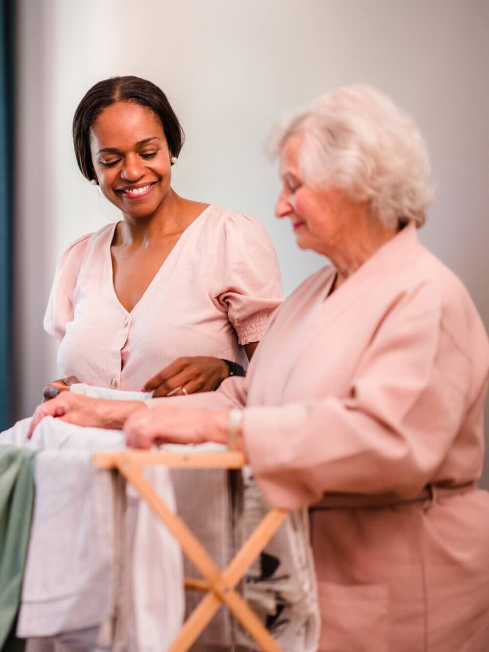 A woman helps an older lady with laundry. They both wear light-colored shirts and share a moment of connection. - Home Instead