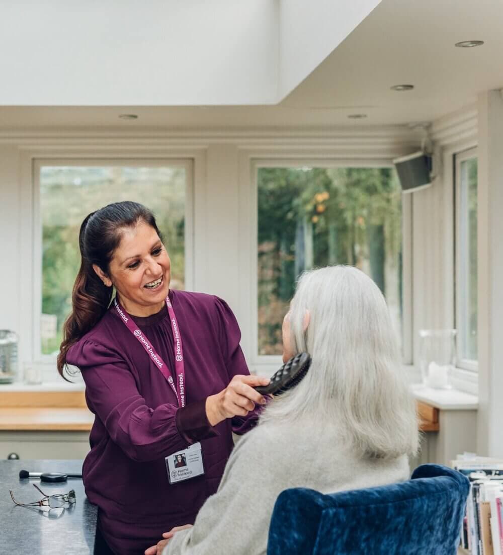 Caregiver combing the hair of a senior man sitting in a wheelchair, both smiling, in a cozy, book-filled room. - Home Instead