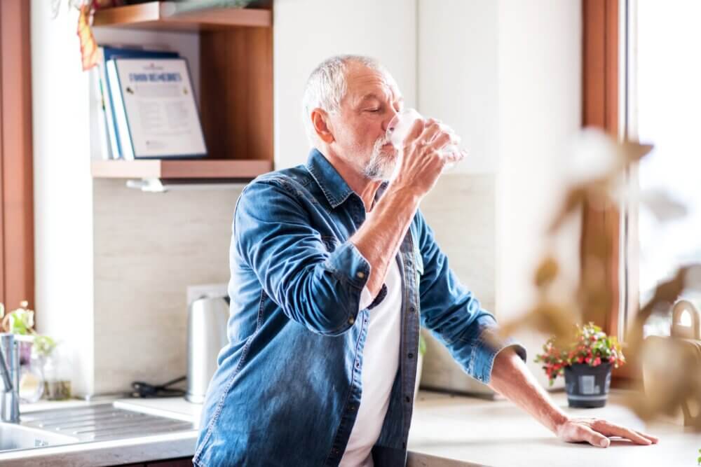 An older man wearing a denim shirt drinks water in a bright kitchen with plants and books in the background. - Home Instead