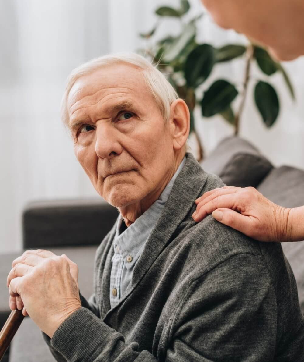 Elderly man sitting and holding a cane while another person places a reassuring hand on his shoulder. - Home Instead