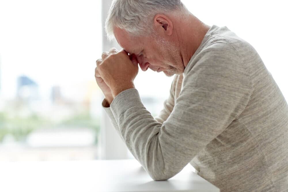 Elderly man in a sweater, sitting at a table, resting his face on his clasped hands, appearing contemplative or stressed. - Home Instead