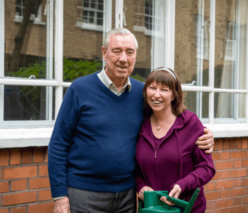 Older man and woman smiling, standing together in front of a window. The woman is holding a green watering can. - Home Instead