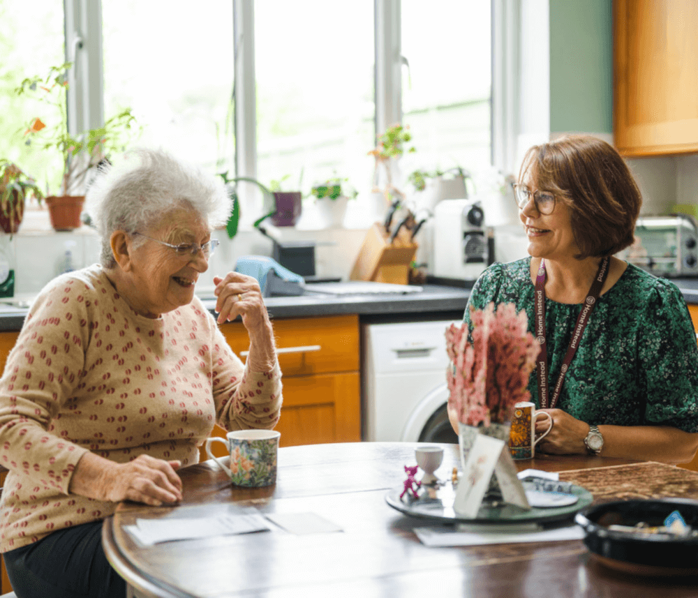 An elderly woman and a younger woman talking and smiling while seated at a kitchen table with mugs. - Home Instead