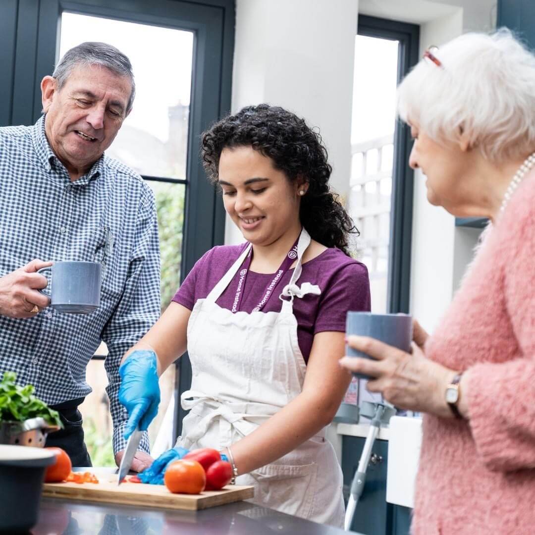 Young woman chopping vegetables in a kitchen while an elderly man and woman hold mugs and watch. - Home Instead
