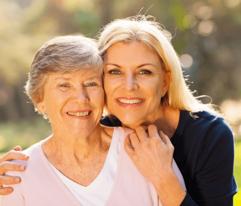 Two smiling women, an older woman with short gray hair and a younger woman with long blonde hair, hugging in a sunny outdoor setting. - Home Instead