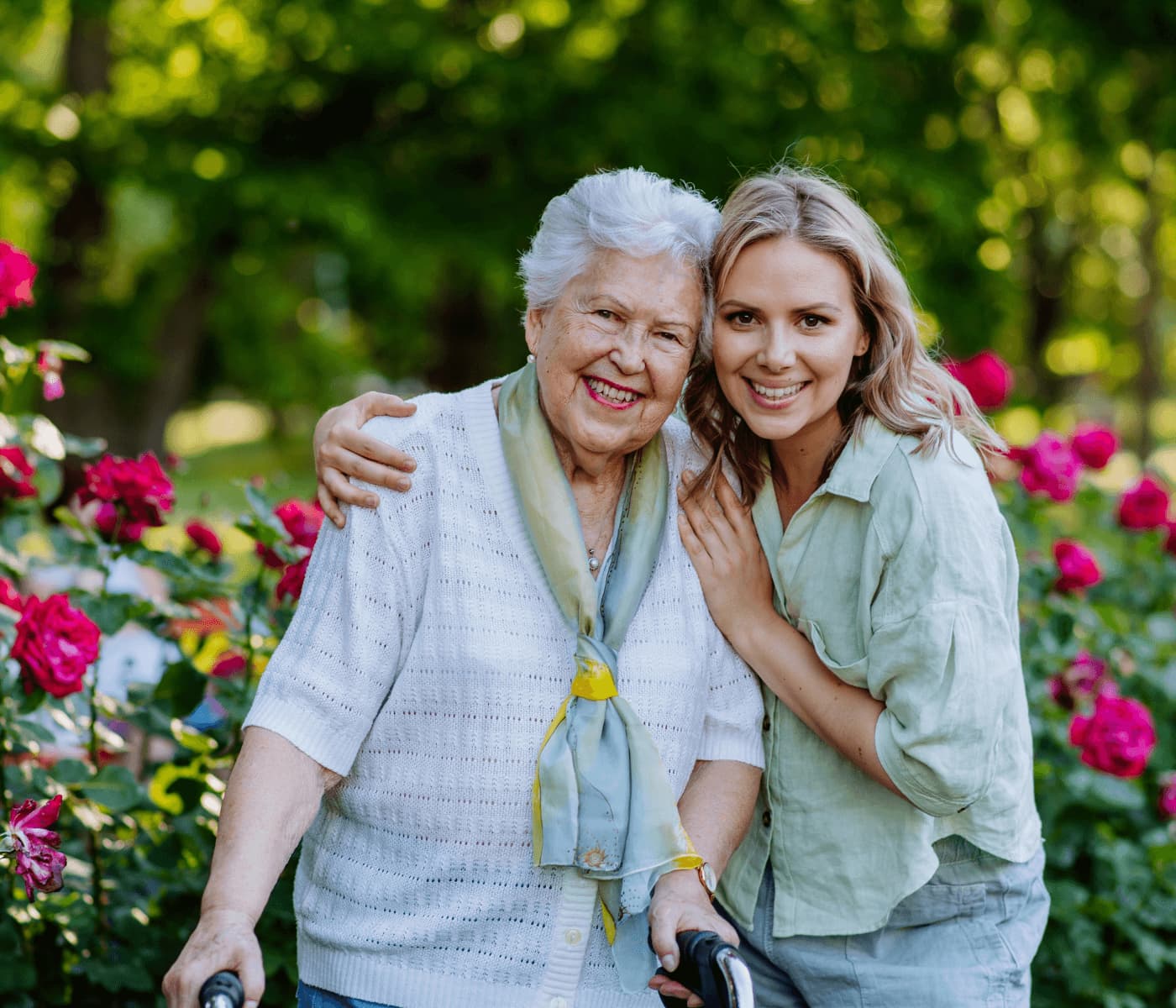 An elderly woman with a walker and a young woman smile and pose together in a garden with blooming rose bushes. - Home Instead