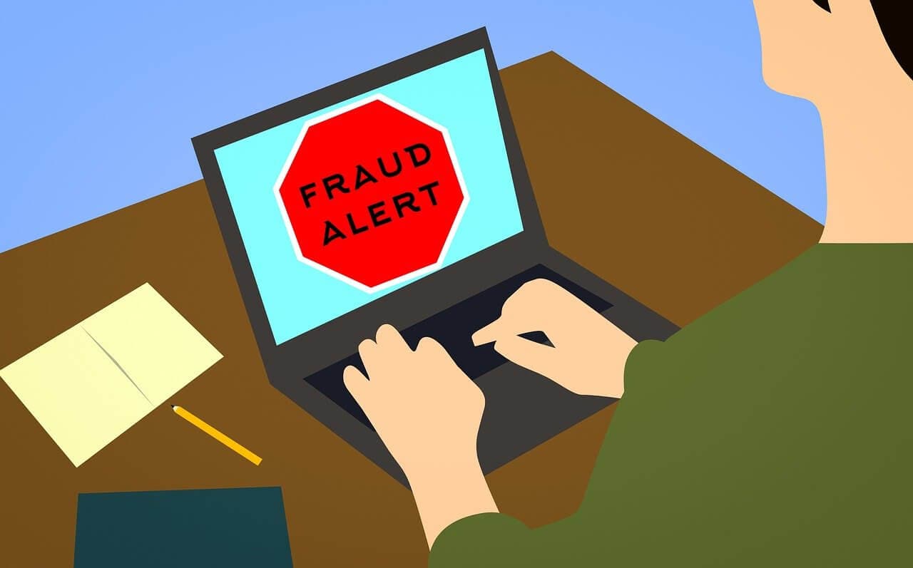 Person using a laptop displaying a red stop sign with "FRAUD ALERT". A pencil and papers are on the table. - Home Instead
