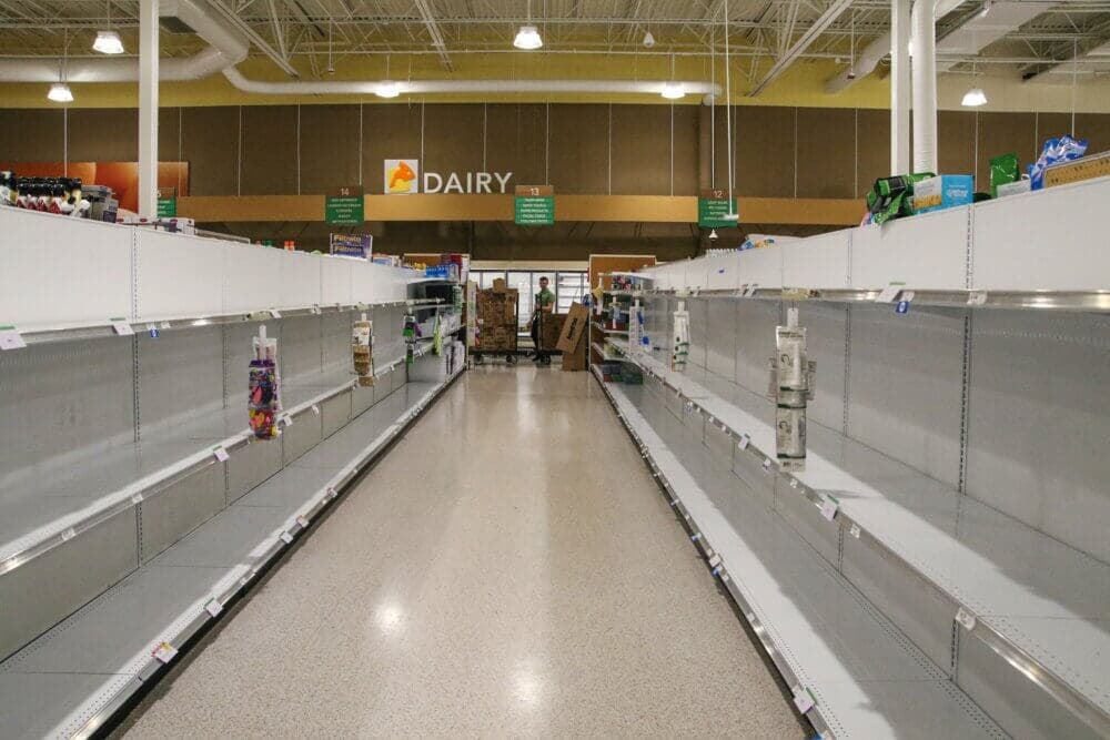 Empty supermarket shelves with a few remaining items near the dairy section, overhead lights illuminate the scene. - Home Instead
