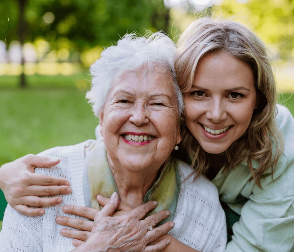 An elderly woman and a younger woman smiling and hugging outdoors. - Home Instead
