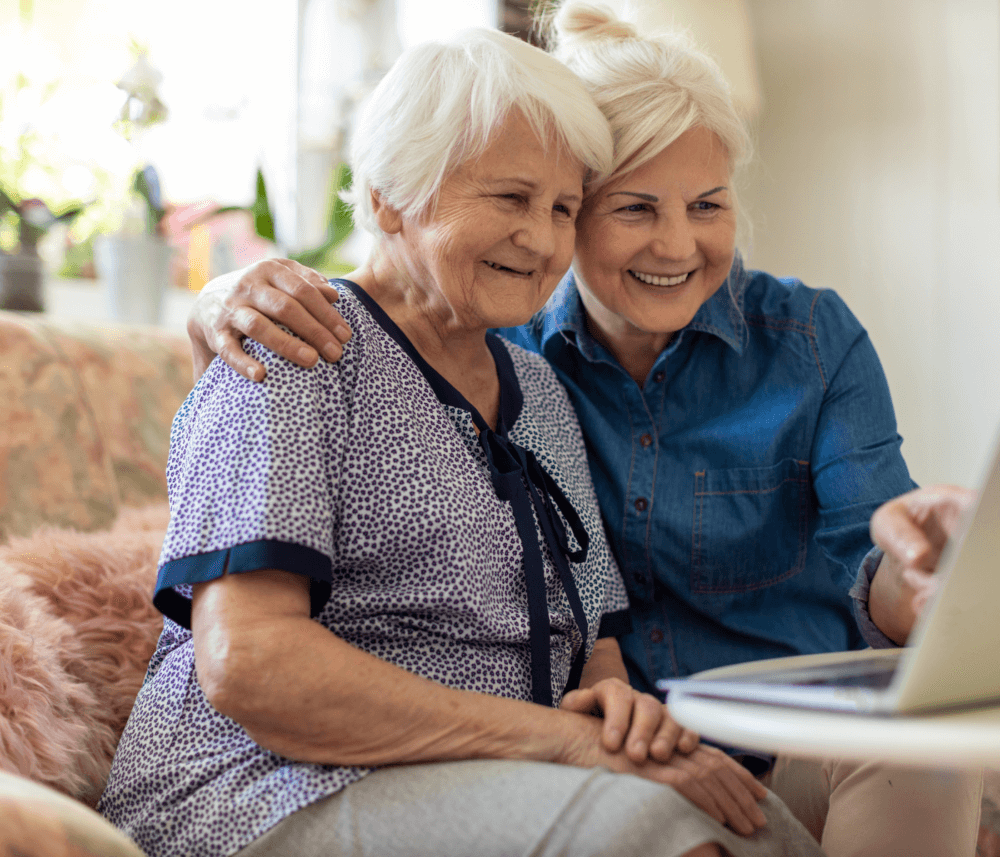 Two smiling elderly women sitting on a couch, looking at a laptop together, with one arm around each other. - Home Instead