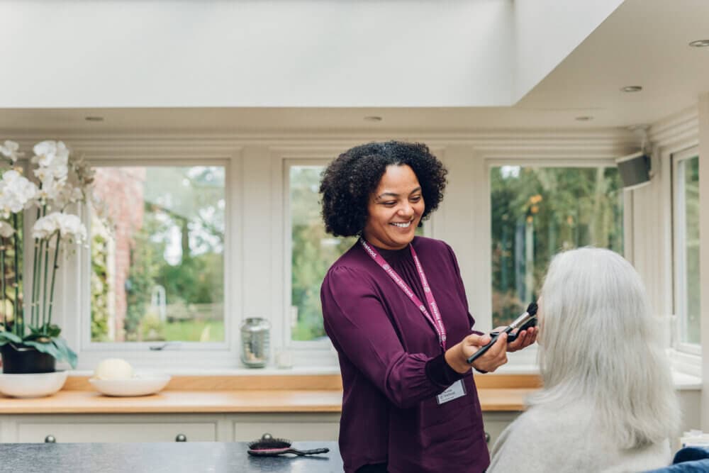 A caregiver with a name tag helps an elderly woman brush her hair in a bright, modern kitchen. - Home Instead
