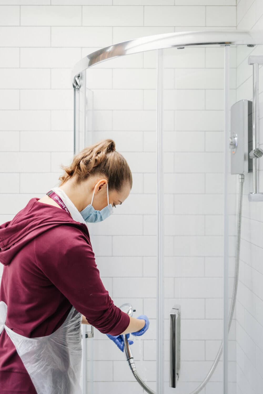 Person cleaning a glass shower door while wearing a mask, gloves, and apron in a bathroom with white tiled walls. - Home Instead