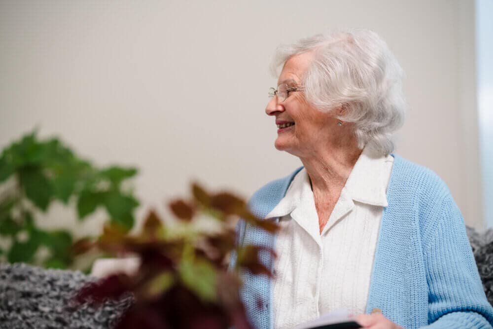 Elderly woman with white hair and glasses smiles while sitting indoors, wearing a blue cardigan over a white blouse. - Home Instead