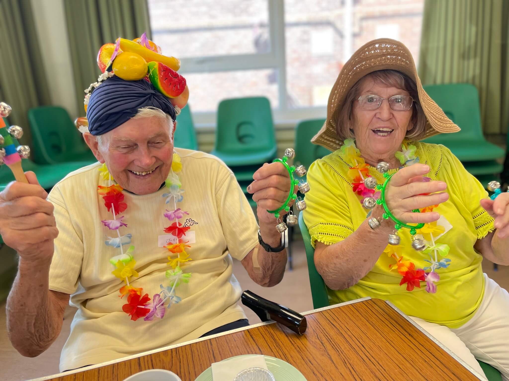 Two elderly individuals wearing colorful attire and hats, joyfully shaking tambourines at a table. - Home Instead