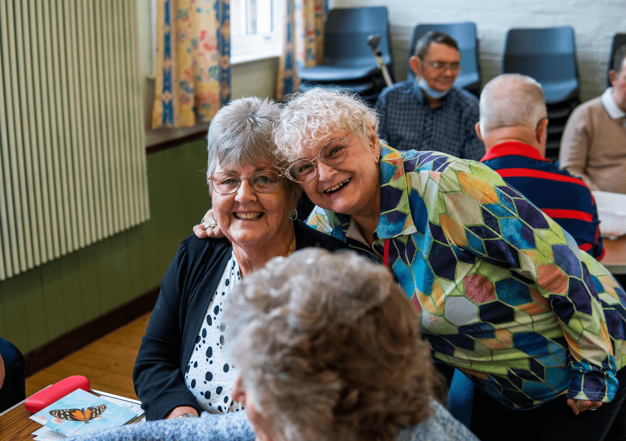 Two smiling elderly women pose together at a community gathering, with others chatting in the background. - Home Instead