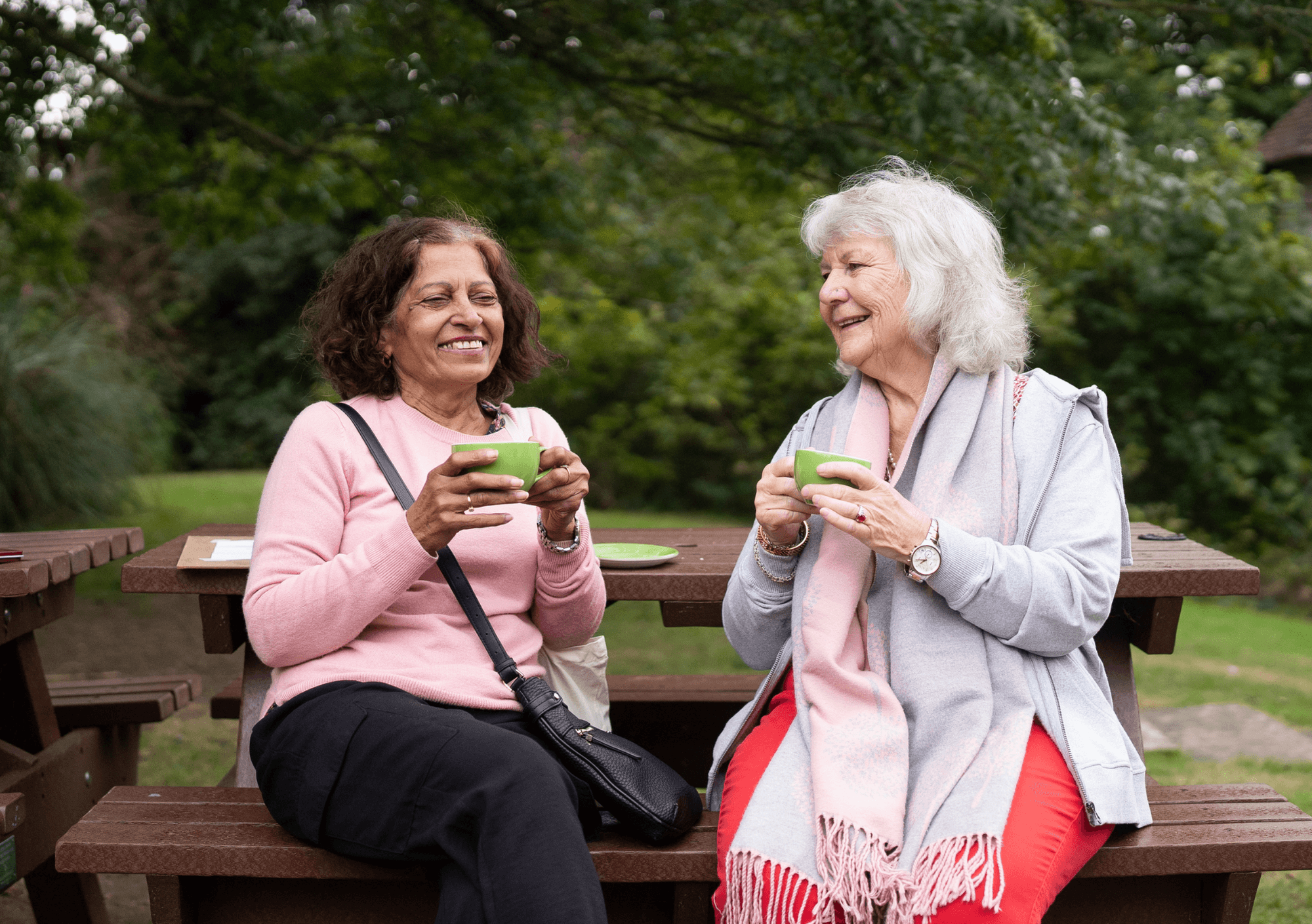 Two older women sit on a bench outdoors, smiling and holding green mugs, with trees in the background. - Home Instead